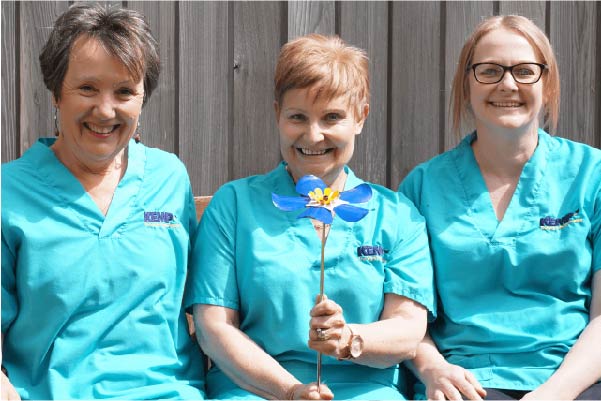 Three kemp hospice nurses in teal scrubs holding a remembrance blue flower